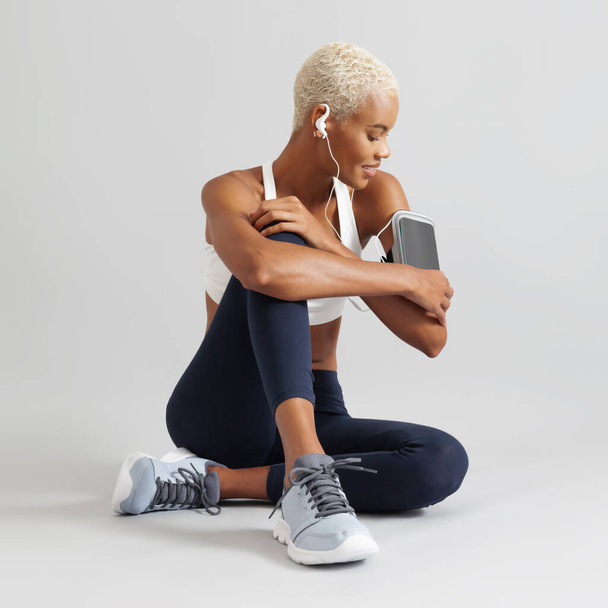 Fitness cvičení, sedící žena se sluchátky dotýkající se obrazovky mobilního telefonu na rameni, latinskoamerická atletka ve sportovním oblečení připravující se na běh, cvičení v posilovně nebo jogging - Fotografie, Obrázek