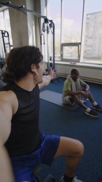 Pionowe ujęcie POV sportowca dzwoniącego do kogoś na wideo i mówiącego o jego przyjacielu robiącym loki na podłodze podczas treningu na siłowni - Materiał filmowy, wideo