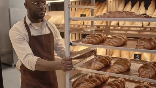 Lento medio del joven vendedor de panadería afroamericana calva en delantal llevando estante de bandeja de aluminio con productos horneados frescos a lo largo de la panadería - Metraje, vídeo