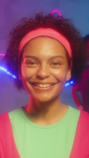 Retrato vertical de mulher afro-americana alegre em activewear retro olhando para a câmera após a aula de fitness no estúdio de néon roxo - Filmagem, Vídeo