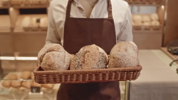 Retrato de slow mo recortado de un trabajador de panadería irreconocible en delantal sosteniendo una bandeja de paja con panes redondos de pan recién hechos, de pie en una pequeña panadería acogedora - Metraje, vídeo