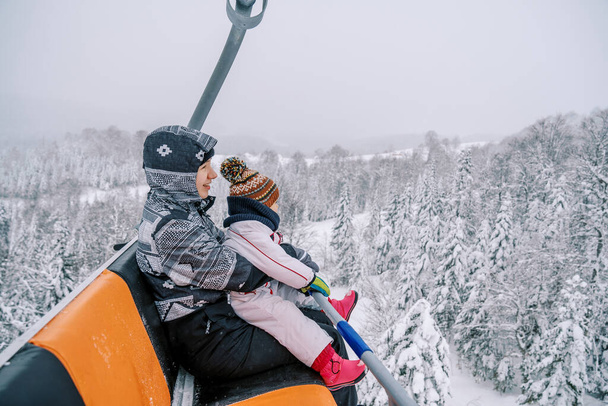 Äiti, jolla on pieni lapsi sylissään hiihtopuvuissa, nousee vuorelle tuolihissillä. Laadukas kuva - Valokuva, kuva