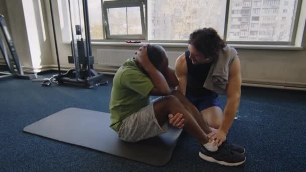 Χειροκίνητη βολή του ανδρικού προπονητή βοηθώντας τον πελάτη άσκηση στο γυμναστήριο και να κάνει κοιλιακή crunches στο πάτωμα - Πλάνα, βίντεο