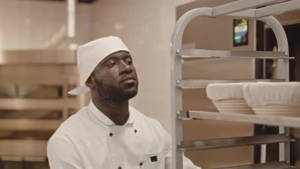 Cintura para arriba de un joven trabajador de panadería afroamericano en uniforme de chef llevando bandeja con cestas de pan a lo largo de la cocina y tomando el plan diario en hoja de papel de refrigerador de acero inoxidable - Imágenes, Vídeo
