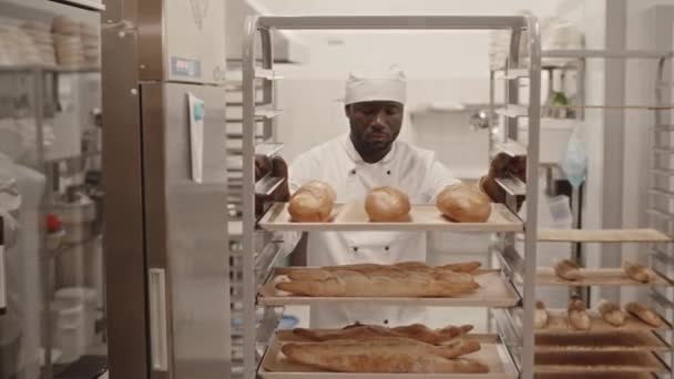 Lentidão média de padeiro macho preto em uniforme de chef transportando rack de bandeja de alumínio com pães frescos e baguetes ao longo da cozinha da padaria - Filmagem, Vídeo