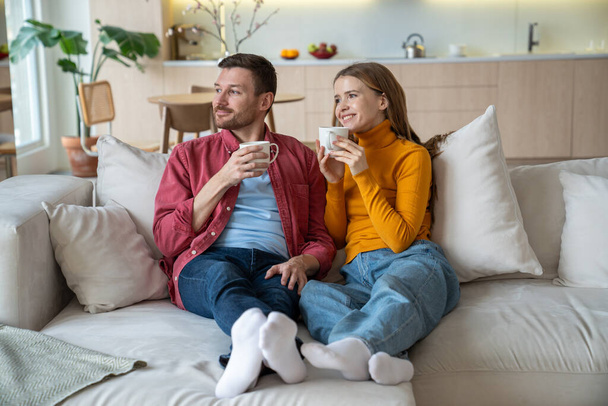 Married couple spending weekends at home together. Happy loving wife and husband sitting on sofa, drinking tea, looking at window, dreaming of children. Tenderness, closeness in young family life - Photo, Image