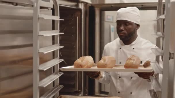 Μέτριο πλάνο του Αφροαμερικανού εργάτη αρτοποιίας με στολή λευκού σεφ λαμβάνοντας ταψί με φρέσκα καρβέλια ψωμί εκτός φούρνου και βάζοντας τα σε σχάρα δίσκου - Πλάνα, βίντεο