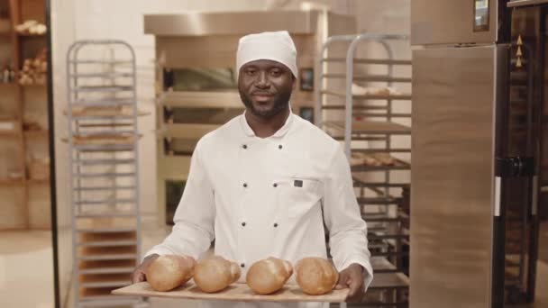 Medium slow mo portret van Afro-Amerikaanse mannelijke bakker in chef-kok uniform poseren voor camera met net gemaakte broden op houten dienblad in bakkerij keuken - Video