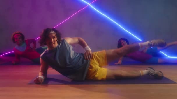 Male coach and two biracial sportswomen doing side leg raises on floor in retro outfits - Footage, Video