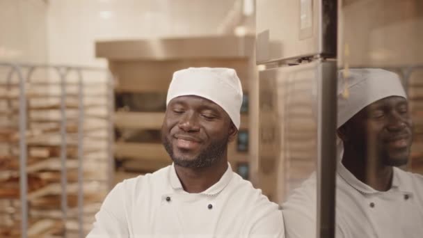 Cintura hacia arriba retrato slow mo de un chef negro posando con las manos dobladas en la cámara mientras se apoya en el refrigerador en la cocina de panadería - Metraje, vídeo