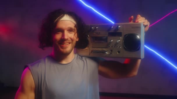 Retrato de deportista masculino alegre en ropa deportiva retro posando para cámara con boombox en hombro en estudio con luz de neón - Metraje, vídeo