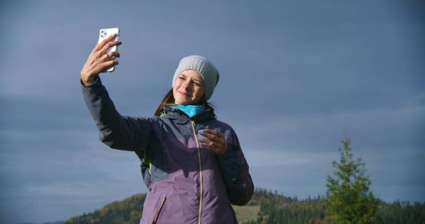 Senderista toma selfie utilizando el teléfono móvil de pie delante de hermosos paisajes en la colina. Mujer caucásica sostiene la taza de té y mira el teléfono inteligente. Turista durante el viaje o trekking en las montañas. - Foto, Imagen