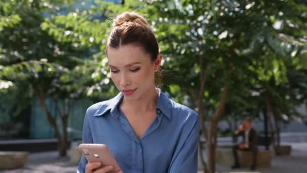 Κορίτσι freelancer βλέποντας smartphone social media κάθεται ηλιόλουστο δρόμο κοντά. Ελκυστική ήρεμη γυναίκα που ψάχνει κινητό τηλέφωνο περιμένει συνάντηση στο πάρκο. Αυτοπεποίθηση νεαρή κυρία ανάγνωση τηλεφωνικό μήνυμα εξωτερική. - Πλάνα, βίντεο