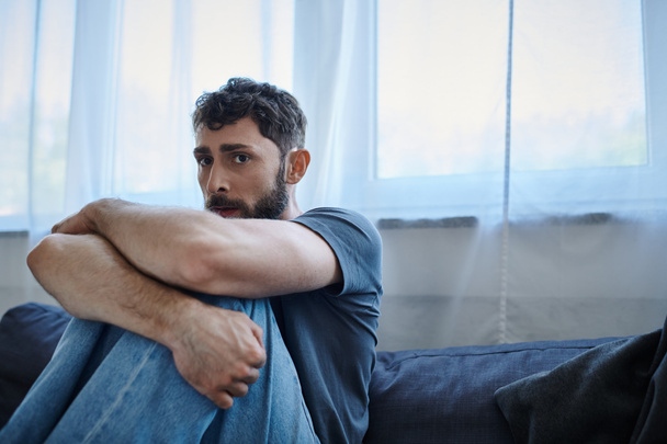 homme anxieux barbu en tenue décontractée assis sur le canapé pendant la dépression mentale et en regardant la caméra - Photo, image
