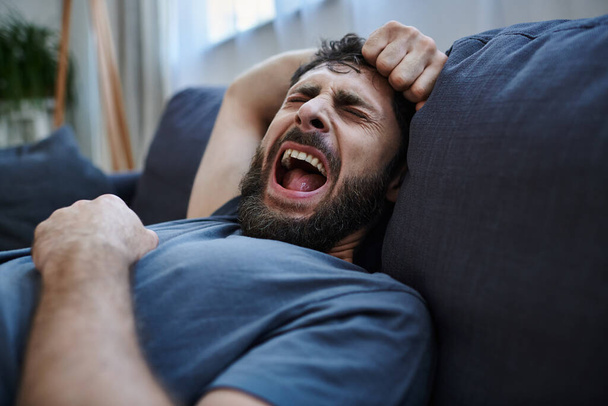 zdesperowany mężczyzna w depresji w homewear na kanapie krzyczy podczas załamania, świadomości zdrowia psychicznego - Zdjęcie, obraz