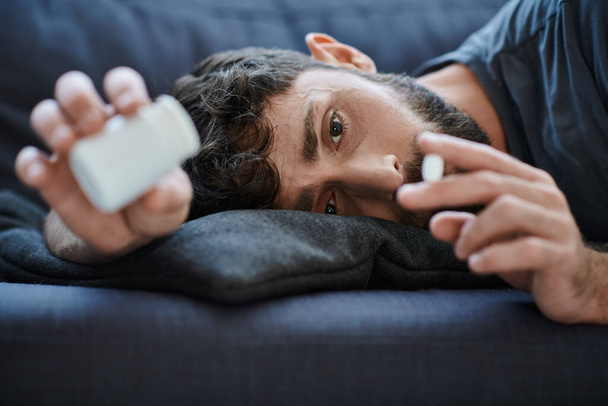 homem sofredor em traje casual deitado no sofá com pílulas na mão, consciência de saúde mental - Foto, Imagem