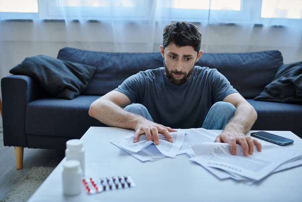 depresivní nemocný muž s vousy sedí u stolu s papíry a prášky na něm během depresivní epizody - Fotografie, Obrázek