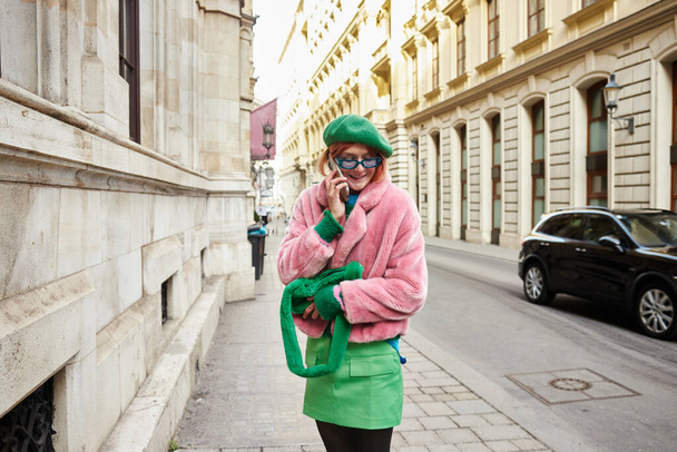 Sahte kürk ceketli, güneş gözlüklü, gülümseyen şık bir kadın Viyana 'da sokakta akıllı telefondan konuşuyor. - Fotoğraf, Görsel