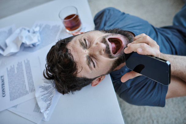 frustrado hombre gritando en el teléfono inteligente con un vaso de alcohol a su lado durante la crisis mental - Foto, imagen