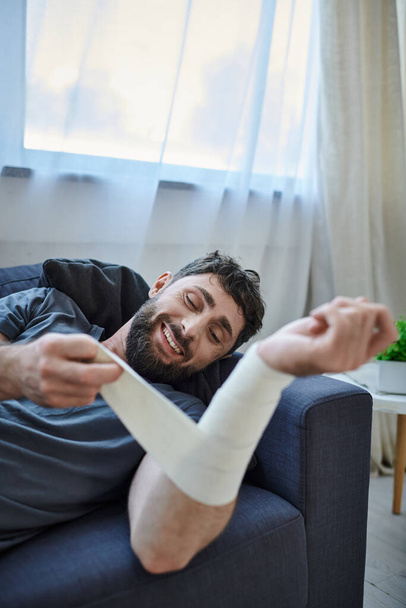 hombre enfermo con sonrisa y vendaje en el brazo después de intentar suicidarse en el sofá, conciencia de salud mental - Foto, imagen