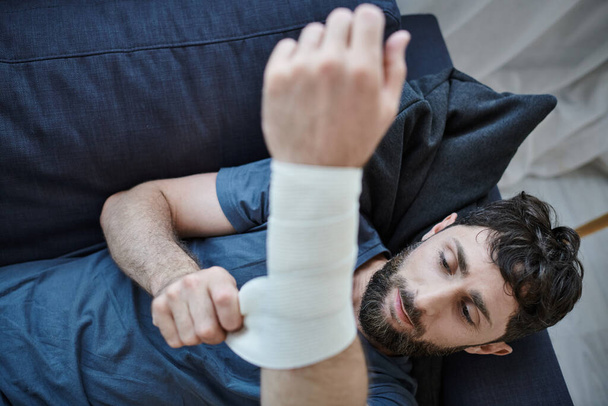 traumatizált férfi kötéssel a karján, miután megpróbált öngyilkos lenni a kanapén fekve, mentális egészség tudatosság - Fotó, kép