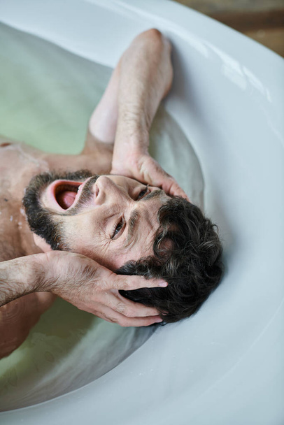 эмоционально травмированный человек, лежащий в ванной и кричащий во время нервного срыва, осознание психического здоровья - Фото, изображение