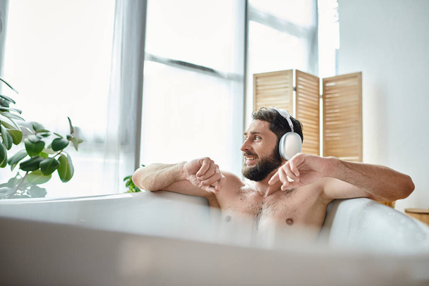 radosny atrakcyjny mężczyzna z brodą i słuchawkami siedzi i relaksuje się w wannie, zdrowie psychiczne - Zdjęcie, obraz