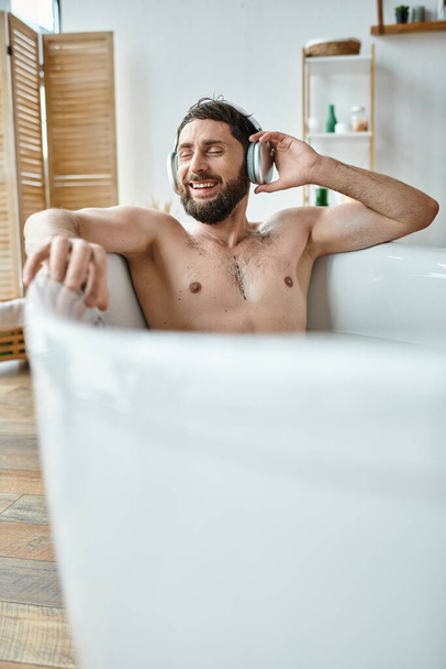 веселый привлекательный мужчина с бородой и наушниками сидит и расслабляется в ванной, психическое здоровье - Фото, изображение