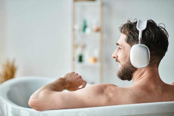 fröhlicher gutaussehender Mann mit Bart und Kopfhörer, der in seiner Badewanne sitzt und entspannt, psychische Gesundheit - Foto, Bild