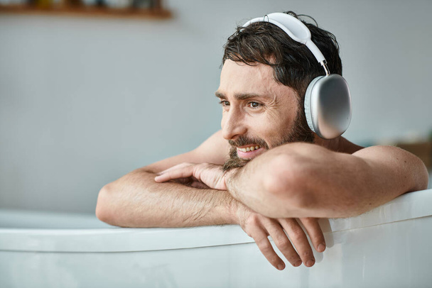 веселый красивый мужчина с бородой и наушниками сидит и расслабляется в ванной, психическое здоровье - Фото, изображение