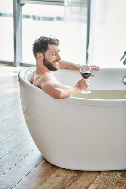 gioioso bell'uomo con barba rilassante nella vasca da bagno con bicchiere di vino rosso, consapevolezza della salute mentale - Foto, immagini