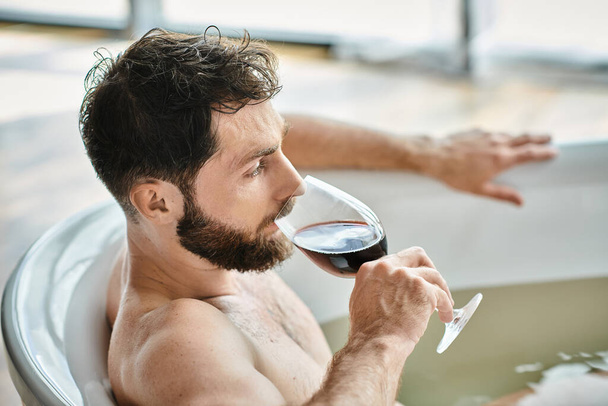 χαρωπός όμορφος άνδρας με γενειάδα χαλαρώνει στην μπανιέρα με ένα ποτήρι κόκκινο κρασί, ψυχική υγεία συνείδηση - Φωτογραφία, εικόνα