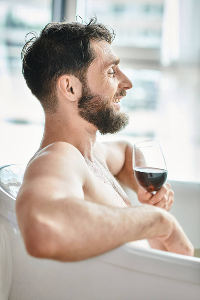 gelukkige aantrekkelijke man met baard ontspannen in bad met een glas rode wijn, geestelijke gezondheid bewustzijn - Foto, afbeelding