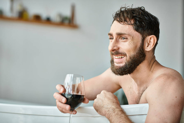 Mutlu, sakallı, küvette bir bardak kırmızı şarapla rahatlayan, zihinsel sağlığın farkında olan çekici bir adam. - Fotoğraf, Görsel