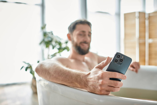 se centran en el teléfono inteligente moderno en manos del hombre barbudo alegre borrosa tomando baño, salud mental - Foto, Imagen