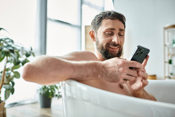 веселый красивый мужчина с бородой, смотрящий на свой смартфон в ванной, психическое здоровье - Фото, изображение