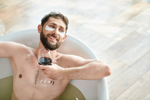 gioioso uomo attraente con barba e macchie oculari rilassante nella vasca da bagno con bicchiere di vino rosso - Foto, immagini