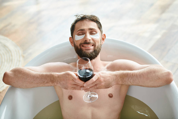 gioioso uomo attraente con barba e macchie oculari rilassante nella vasca da bagno con bicchiere di vino rosso - Foto, immagini