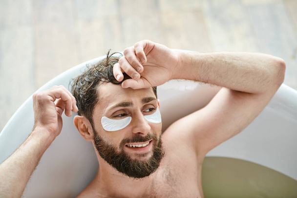весёлый красивый мужчина с бородой расслабляется в ванной с пятнами на глазах, психическим здоровьем - Фото, изображение