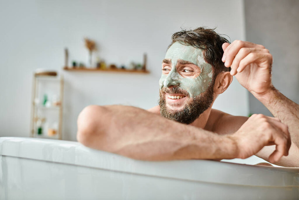 радісний привабливий чоловік з бородою і маскою для обличчя, що охолоджується у ванні, обізнаність про психічне здоров'я - Фото, зображення