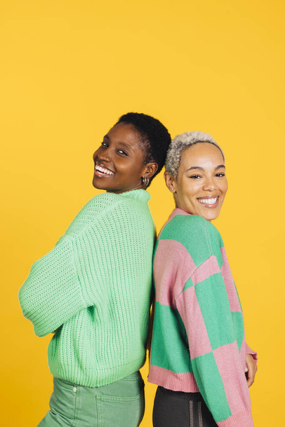 Two young cheerful women wearing green clothes embracing in a studio with a yellow background. - Photo, Image