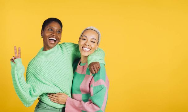 Zwei junge, fröhliche Frauen in grünen Kleidern umarmen sich in einem Atelier mit gelbem Hintergrund. - Foto, Bild