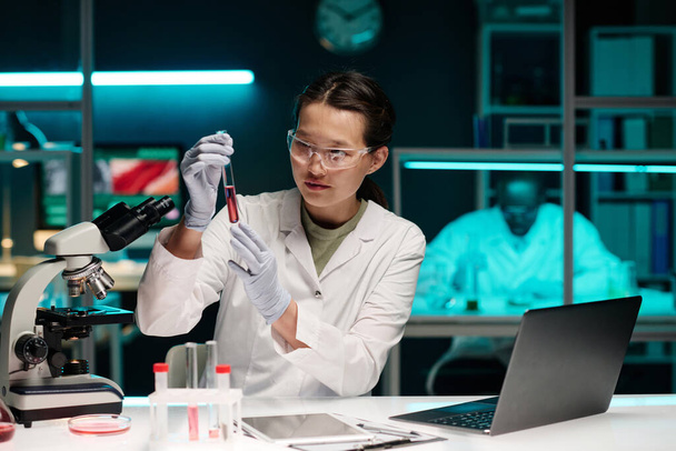 Nuori aasialainen naispuolinen tiedemies katselee vaaleanpunaista nestettä koeputkessa istuu pöydän ääressä laboratoriossa - Valokuva, kuva