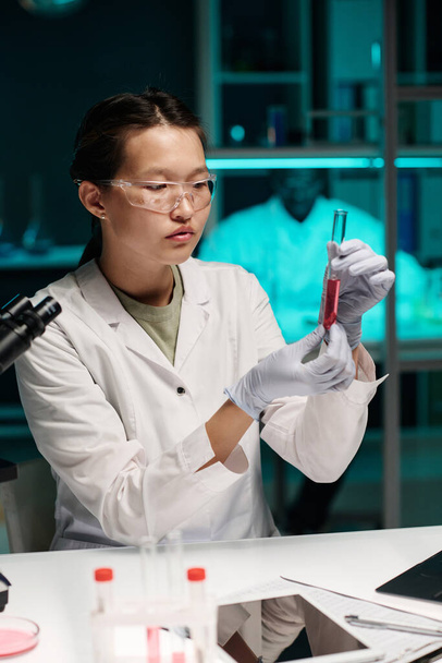 Asiatica scienziata esaminando sostanza rosa in provetta seduta sul suo posto di lavoro in laboratorio - Foto, immagini