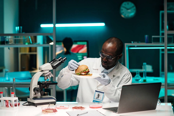 Scienziato afroamericano che esamina hamburger con carne coltivata in laboratorio seduta sul suo posto di lavoro in laboratorio - Foto, immagini