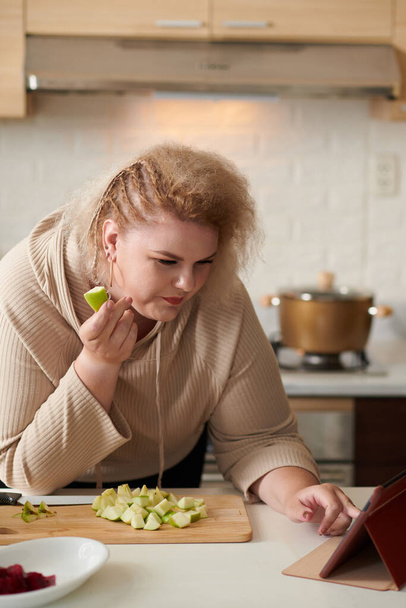 Stylowe plus rozmiar młoda kobieta jedzenie cut apple podczas wyszukiwania receptury wideo na tablecie - Zdjęcie, obraz