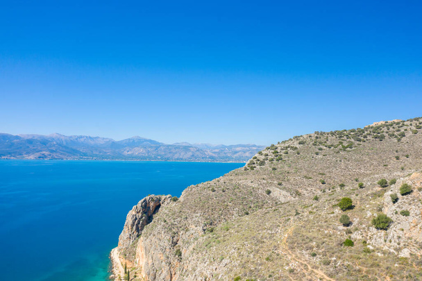 To zdjęcie krajobrazowe zostało zrobione latem w Europie, Grecji, na Peloponezie, w Argolis, w Nafplio, nad morzem Myrto. Widzimy suche skaliste wybrzeże i jego zieloną okolicę na skraju małych plaż, pod słońcem. - Zdjęcie, obraz
