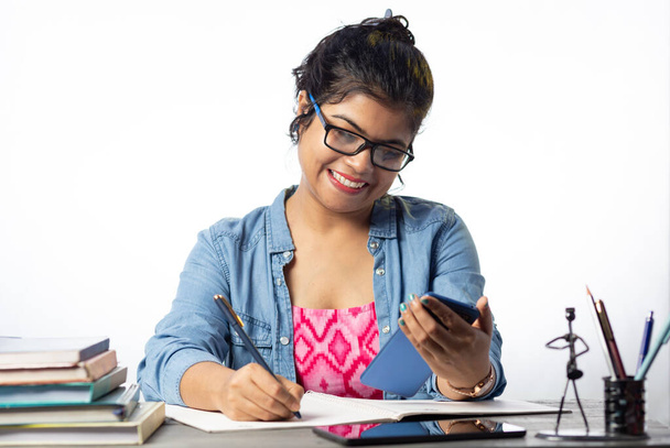 Un joli jeune étudiant indien en lunettes étudiant et écrivant avec téléphone intelligent sur table d'étude et fond blanc - Photo, image