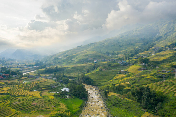 Это пейзажное фото было сделано в Азии, во Вьетнаме, в Тонкине, в Сапе, к Лао Цаю, летом. Мы видим традиционную деревню с ее зелеными и желтыми рисовыми полями в зеленых горах, под облаками. - Фото, изображение