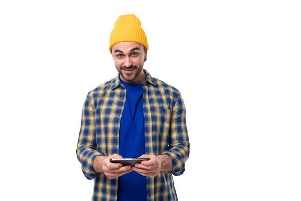 zadbany młody europejski brunetka człowiek z brodą i wąsami ubrany w żółty kapelusz i niebieską koszulę wykorzystuje sieci społeczne w smartfonie. - Zdjęcie, obraz